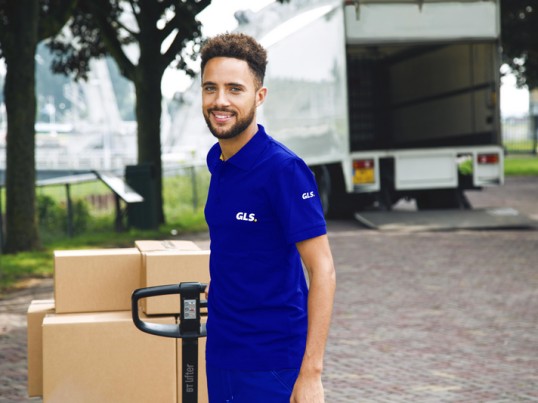 Een GLS-chauffeur in een blauw uniform staat naast opgestapelde kartonnen dozen en een steekwagen voor een open leveringstruck.
