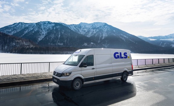 A white GLS delivery van is parked on a wet road beside a railing, with a snowy mountain range and lake in the background.