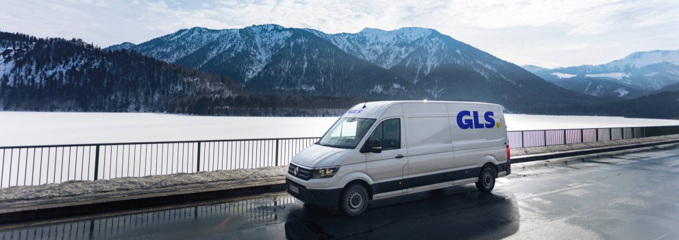 A white GLS delivery van is parked on a wet road beside a railing, with a snowy mountain range and lake in the background.