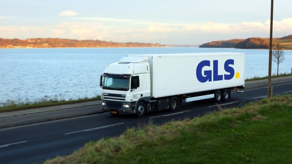 A white GLS delivery truck drives along a lakeside road with hills and water in the background.