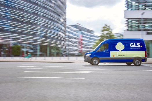 Een blauwe GLS elektrische bezorgbus rijdt over een stadsstraat voor moderne glazen kantoorgebouwen.