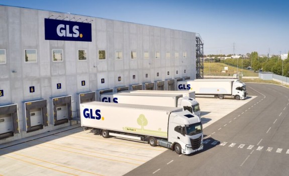 Een groot GLS-logistiek depot met laadperrons en geparkeerde vrachtwagens, gezien van boven, naast verharde wegen en grasvelden.